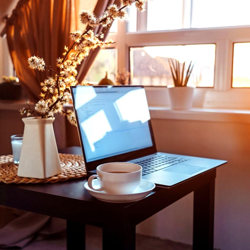 A cozy and inviting workspace bathed in warm natural light. A laptop sits on a dark wooden table near a window, accompanied by a white ceramic cup of coffee and a minimalist vase with delicate dried flowers. The soft golden glow highlights the peaceful atmosphere, creating a perfect setting for creativity and deep focus.